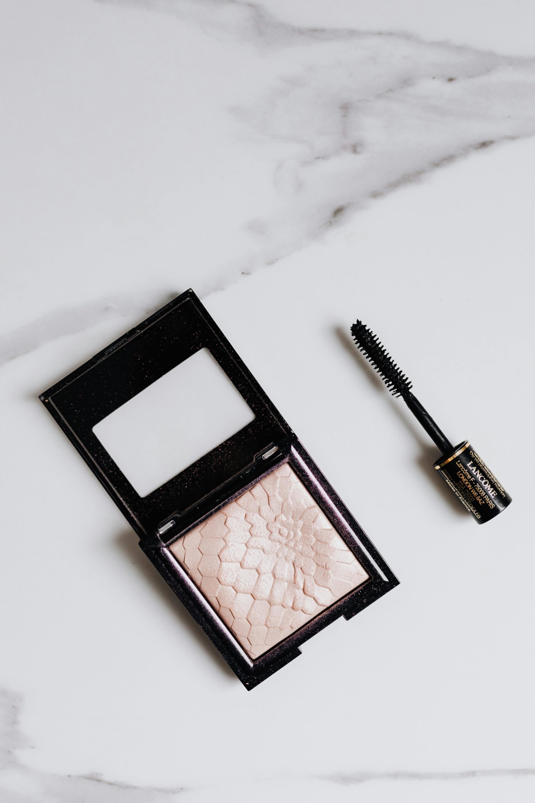 Flat lay of mascara and highlighter compact on a marble background.